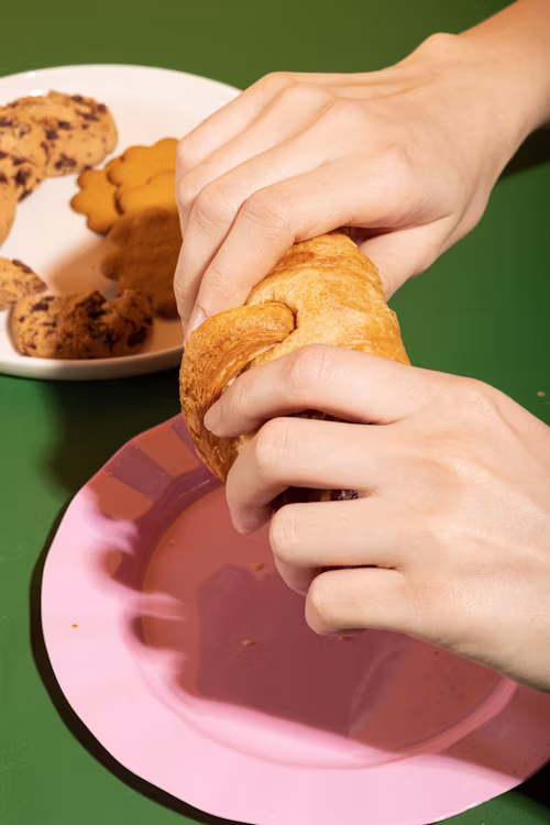 Flaky pastry being torn in half with visible layers, warm natural light, crumbs on a plate, candid hands, shallow depth of field